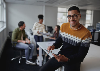 Poster - Portrait of a successful smiling businessman holding digital tablet looking at camera sitting in front of team at modern office