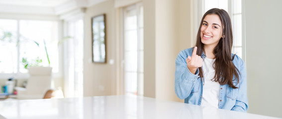 Poster - Wide angle picture of beautiful young woman sitting on white table at home Beckoning come here gesture with hand inviting happy and smiling