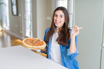 Poster - Beautiful young woman eating homemade tasty pizza at the kitchen very happy pointing with hand and finger to the side