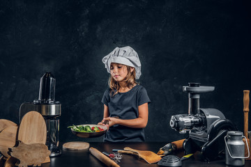 Wall Mural - Cute little girl in chef's hat is preparing vegetables for cooking on the dark background.