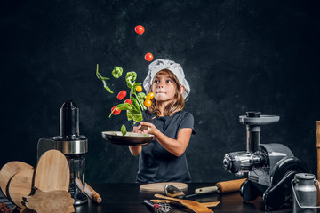 Wall Mural - Pretty little girl is tossing vegetables on the pan at dark photo studio.