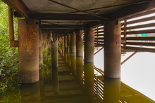 Rusty Metal Columns Of The Base Of A Wooden Pier In The Water Of A River In Summer. View Under The Pier.