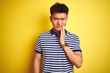 © Krakenimages.com - Young asian chinese man wearing striped polo standing over isolated yellow background touching mouth with hand with painful expression because of toothache or dental illness on teeth. Dentist concept.