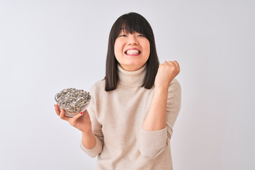 Wall Mural - Beautiful chinese woman holding bowl with sunflowers seeds over isolated white background screaming proud and celebrating victory and success very excited, cheering emotion