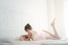 Selective Focus Rear View Of Happy Young Little Girl In Pink Ballet Dress Lying Down On White Bed In Bedroom Alone. Cheerful Pretty Ballerina Child Girl With Playing Mini Red Heart In Gift Box