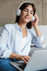 Wall Mural - Relaxing woman indoors at home sit on sofa in living room listening music with headphones using laptop computer.