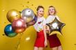 © Вячеслав Косько - Two happy beautiful woman friend celebrating posing surrounded by colorful air balloon confetti. Attractive smiling female wearing bright clothes enjoying party isolated at yellow studio background