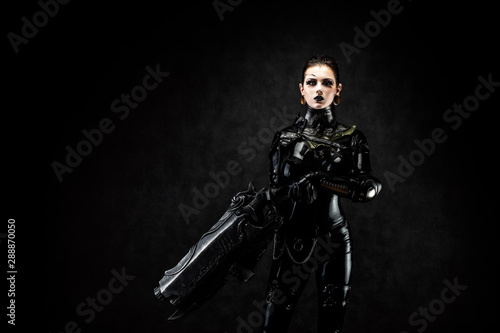 Portrait of a young woman in latex jumpsuit holding a big futuristic weapon in her hands