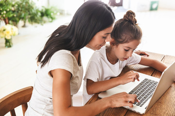 Wall Mural - Image of pretty family woman and her little daughter smiling and using laptop computer together in apartment