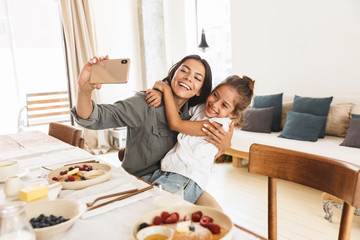 Wall Mural - image of smiling family mother and little daughter taking selfie photo while having breakfast at hom