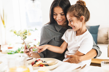 Wall Mural - Image of happy family mother and little daughter looking at cellphone while having breakfast at home in morning