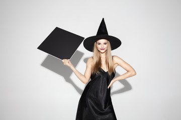 Wall Mural - Young blonde woman in black hat and costume on white background. Attractive caucasian female model. Halloween, black friday, cyber monday, sales, autumn concept. Copyspace. Holding black copyspace.