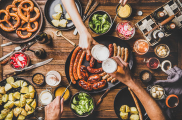 Flat-lay of Oktoberfest dinner table with grilled meat sausages, pretzel pastry, potatoes, cucumber salad, sauces, beers and peoples hands clinking glasses over dark wooden background, top view