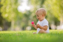 Baby In A White Bodysuit Sitting On The Green Grass Playing