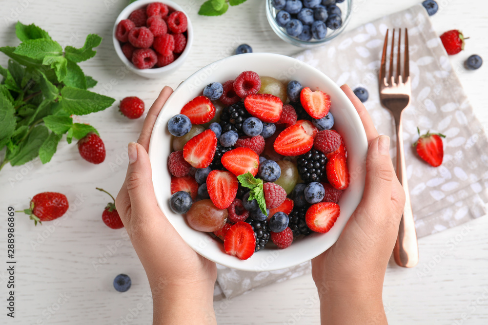 Woman holding bowl with fresh tasty fruit salad over white wooden table, top view - obrazy, fototapety, plakaty 