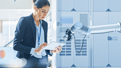 Leinwandbilder - Stylish Female Industrial Robotics Engineer Uses Tablet Computer to Program and Manipulate Robot Arm for Lifting, Moving Objects. Technology Research Facility for Machine Learning and Cloud Computing