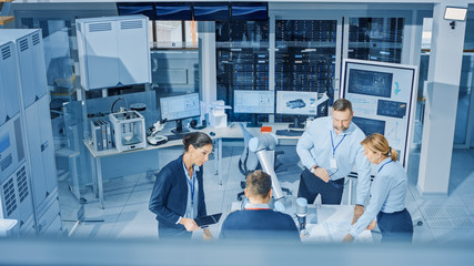 Wall Mural - Diverse Team of Industrial Robotics Engineers Gathered Around Table With Robot Arm, They Manipulate and Program it to Pick Up and Move Metal Component. Bright Facility, Creative People.