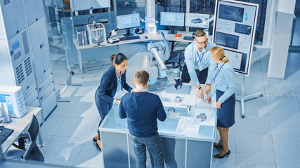 Poster - Diverse Team of Industrial Robotics Engineers Gathered Around Table With Robot Arm, They Manipulate and Program it to Pick Up and Move Metal Component. Bright Facility Creative People. Elevated Camera