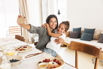 Wall Mural - image of beautiful family mother and little daughter taking selfie photo while having breakfast at h