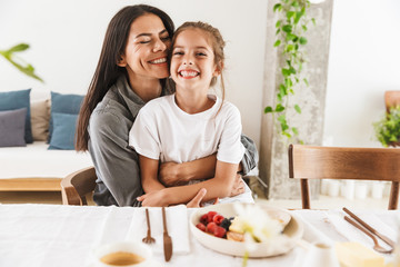 Image of attractive family mother and little daughter hugging while having breakfast at home in morning