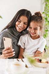 Wall Mural - image of pretty family mother and little daughter looking at cellphone while having breakfast at hom