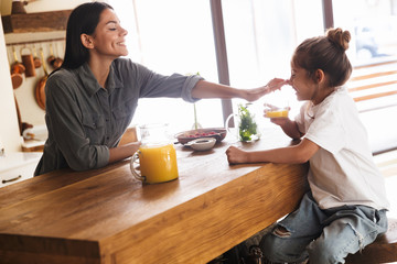 Wall Mural - image of joyous family mother and little daughter drinking orange juice while having breakfast at ho