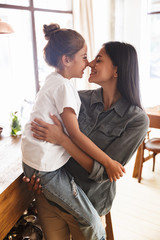 Wall Mural - image of caucasian family mother smiling and holding her little daughter in hands at cozy apartment