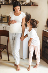 Wall Mural - Full length image of attractive family little girl standing with her pregnant mother at cozy apartment