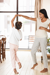 Wall Mural - full length image of joyful family pregnant woman and little girl dancing together at cozy apartment