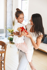 Wall Mural - image of joyful family brunette mother smiling and giving present box to her little daughter at home