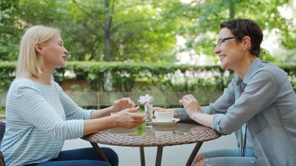 Wall Mural - Mature lady is talking to female friend in open air cafe having fun laughing sitting at table together. Conversation, people and modern lifestyle concept.