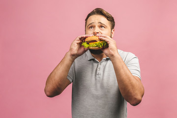 Wall Mural - Young man holding a piece of hamburger. Bearded gyu eats fast food. Burger is not helpful food. Very hungry guy. Diet concept. Isolated over pink background.