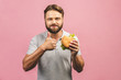 Young man holding a piece of hamburger. Bearded gyu eats fast food. Burger is not helpful food. Very hungry guy. Diet concept. Isolated over pink background. Thumbs up.