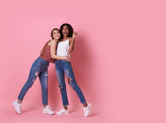 Wall Mural - Portrait of two joyful young woman standing with her finger pointing at copy space isolated over pink banner background.