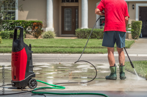 Man using electric powered pressure washer to power wash residential concrete driveway.