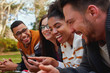 © StratfordProductions - group of multi ethnic friends in college lying together in the park enjoying watching text or video on mobile phone - smiling group of students