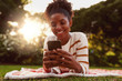 Portrait of a smiling young african woman lying on blanket over green grass texting messages on mobile phone at park