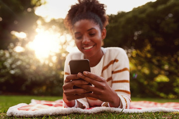 Wall Mural - Portrait of a smiling young african woman lying on blanket over green grass texting messages on mobile phone at park