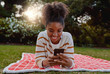 © StratfordProductions - Relaxed smiling young african american woman lying on blanket over lawn in the park using mobile phone - student on their mobile at college park