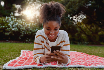 Wall Mural - Relaxed smiling young african american woman lying on blanket over lawn in the park using mobile phone - student on their mobile at college park