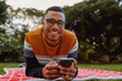 © StratfordProductions - Portrait of a smiling african american young man lying on blanket in the park holding mobile phone in hand looking at camera smiling