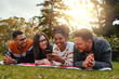 © StratfordProductions - Smiling african american woman lying with her multiethnic group of friends texting on mobile phone in a park - very happy group of friends