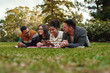 © StratfordProductions - Smiling african american young woman student showing something interesting to her friends lying together on blanket over the green grass in park