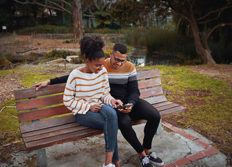 Canvas Print - Happy young african couple sitting together on bench near pond in park using mobile phone