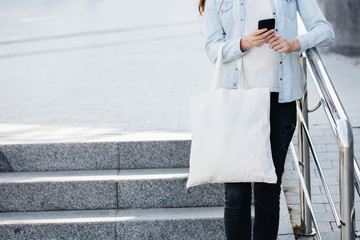 Woman holding white textile eco bag against urban city background. Ecology or environment protection concept. White eco bag for mock up.