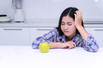 Unhappy young women feeling dislike while looking at green apple on white table in kitchen.