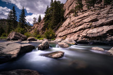 Long Exposure Scene With A River In A Mountain Landscape. The Water Looks Smooth And Silky. The Sky Has Clouds. There Are Forest Pine Trees Visible In The Scene As Well. 