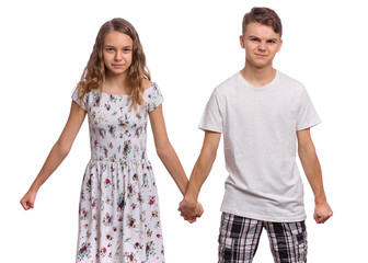 Wall Mural - Portrait of angry teen boy and girl holding hands together. Embittered teenagers looks at camera. Serious children in casual clothes, isolated on white background. Friendship and love concept.