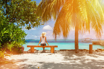 Wall Mural - Fashion woman siting on a bench at the beach. Happy island lifestyle. White sand, blue cloudy sky and crystal sea of tropical beach. Vacation at Paradise. Ocean beach relax, travel to Maldives islands