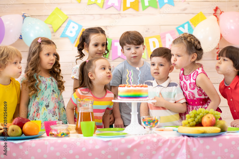Happy children at table celebrating birthday holiday. Kids blows together candles on festive cake - obrazy, fototapety, plakaty 